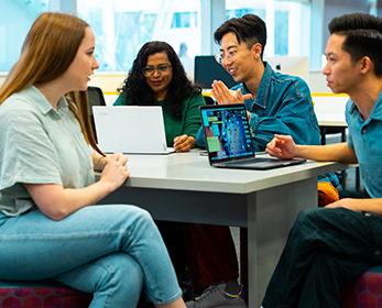 Four students are seated around a table and two discussions are in progress. Two of the students are discussing an item featured on a laptop in front of them.
