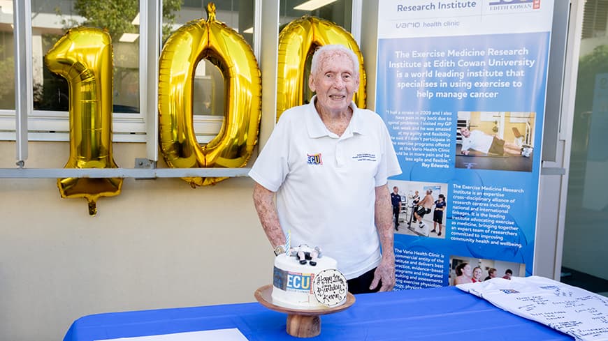 Kevin Ryan who turned 100 years old poses with his birthday cake, there are gold balloons shaped as the number 100 in the background.