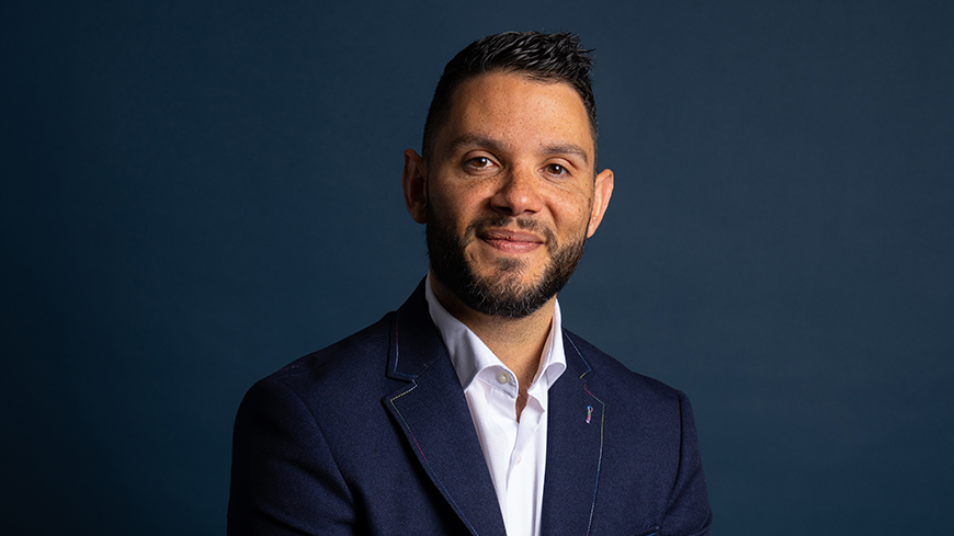 Professor Braden Hill wears a navy suit, has his hands clasped together and sits for his staff profile photo, the backdrop is a dark navy colour.