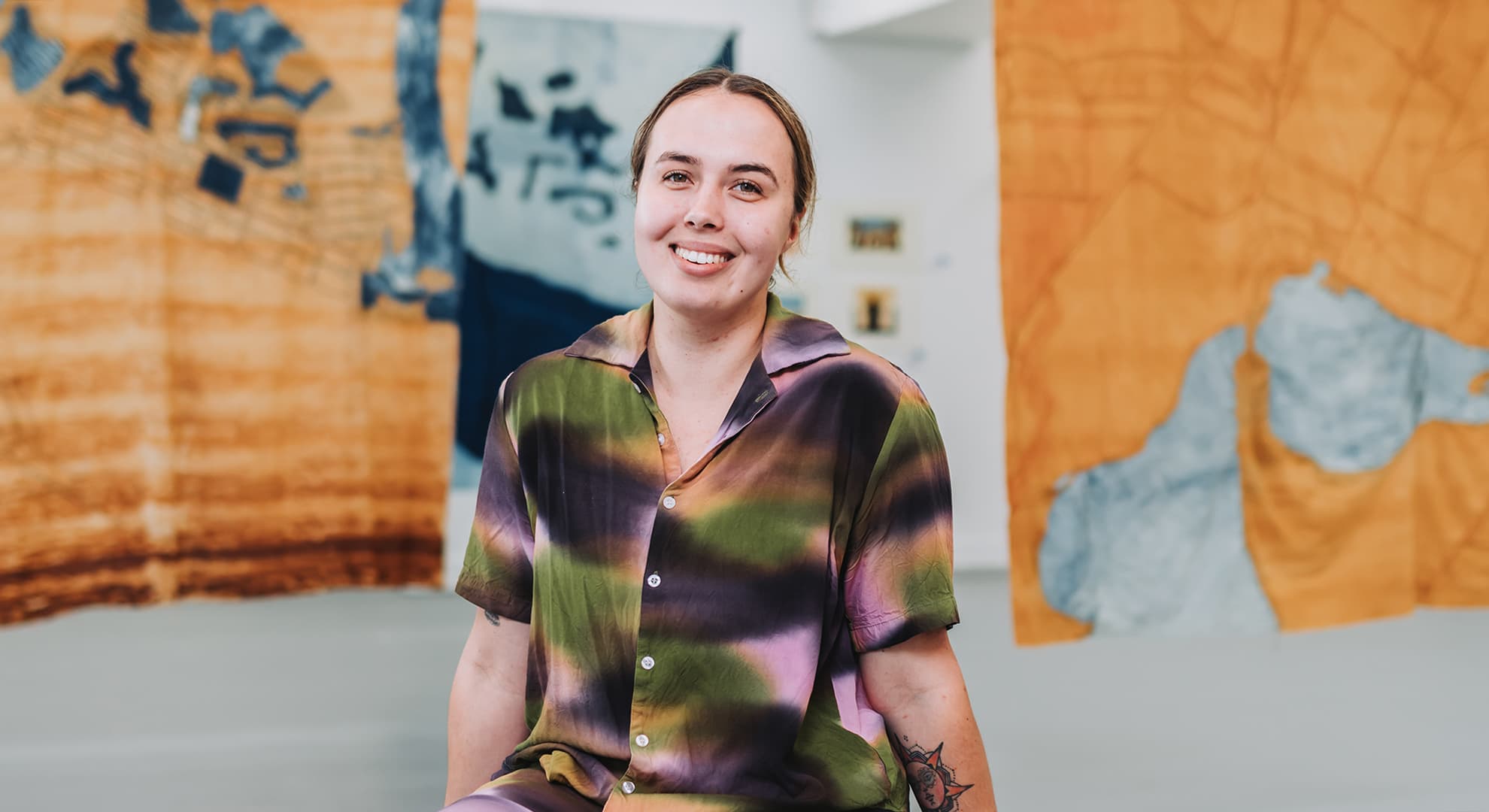 Young person sitting on light grey fabric bench with orche fabric artworks hanging behind her