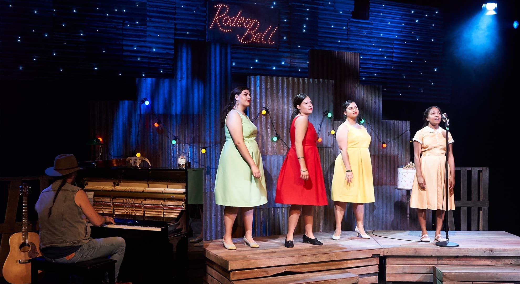 Four women on stage singing, with a man to the side playing piano.