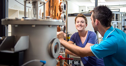 Woman facing camera, man facing away from camera, standing next to marine engineering equipment 