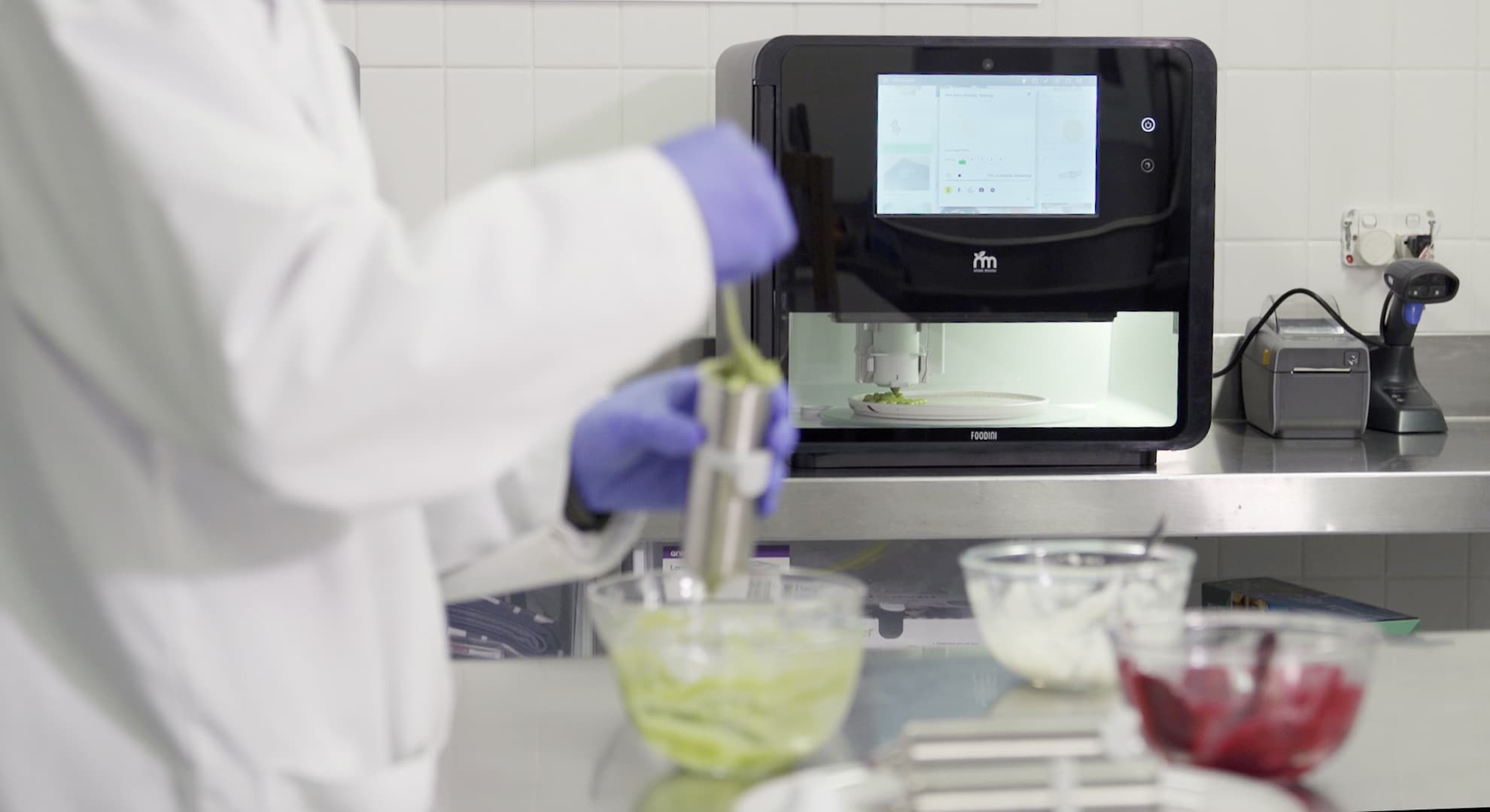 Photo of a lab with person cutting up fruit and vegetables and a 3d printer making a meal in the background.