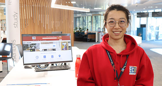A Library Peer Assistant is smiling towards the camera, at the Joondalup Campus Library. She is wearing the red jumper which is the peers' uniform.