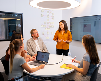 People in a relaxed office setting collaborate on a project together. One person is leading the group through project content displayed on a large wall-mounted screen.