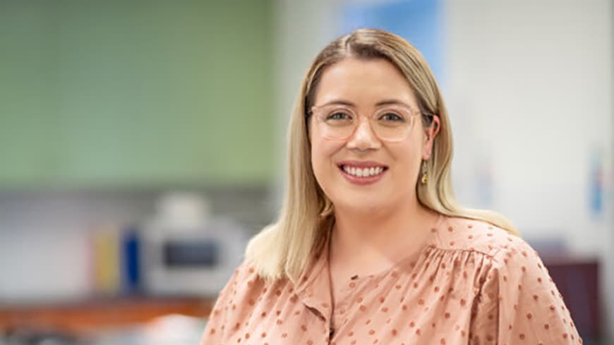 Michaela De Paolis has shoulder-length blonde hair and wears glasses, she is wearing a polka-dot pink blouse and has an ECU lab coat on her arm.