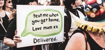 A group of young people gather at a protest. One holds a placard showing a hand-painted text message received that says 'Text me when you get home. Love Mum xx Delivered'. 