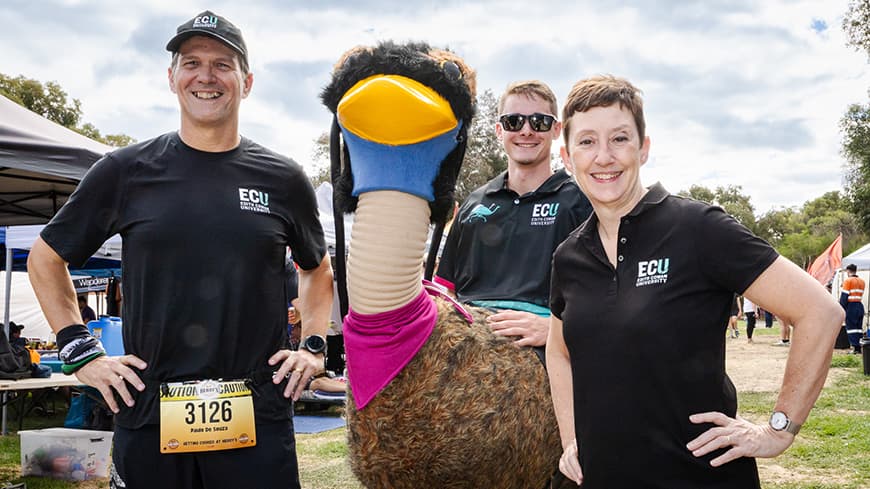 Three people standing in a row with the one in the middle wearing an emu costume
