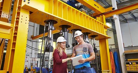 Young man and woman in white hard hats in front of large industrial hoist