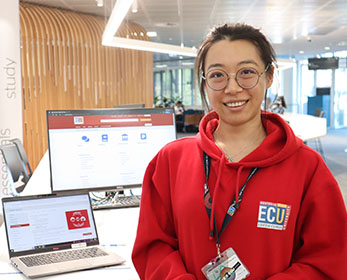A friendly Library Peer Assistant is wearing the red peer uniform. She is smiling at the camera, situated at level 2 of the Joondalup Campus Library with a laptop.