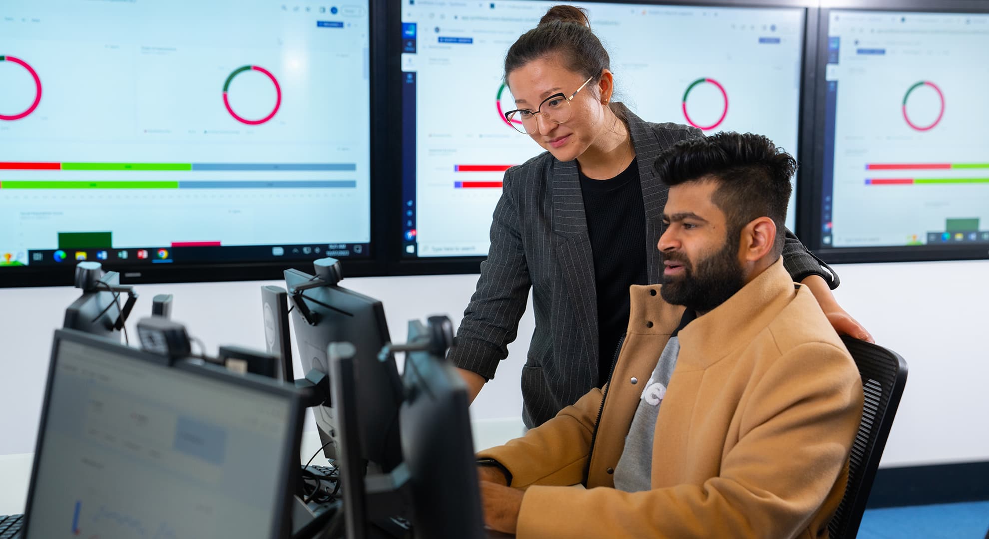 A Master of Business Administration lecturer looks over a student's work on the computer. There are TV screens on the wall displaying graphs.