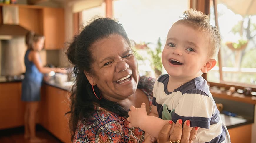 Australian Aboriginal woman holding toddler both smiling close to the camera with girl at sink in blurred background