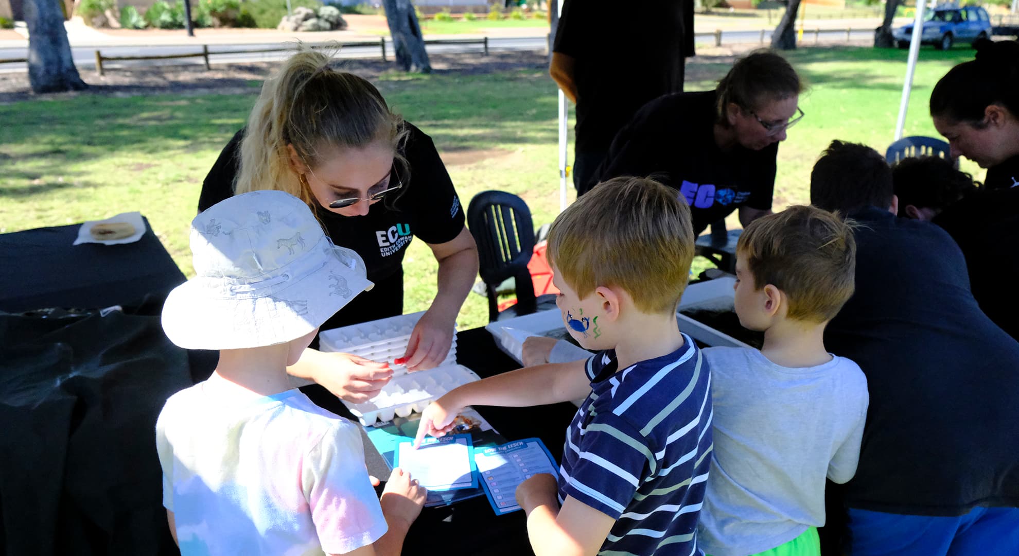 An ECU staff member stands at a stall at a community festival helping participants look for macroinvertebrates in seaweed samples.