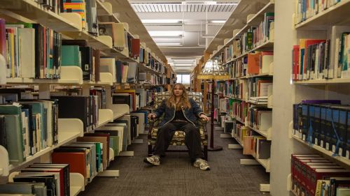 A young adult sitting in an armchair under a lamp in the middle of an aisle in a library, surrounded by books