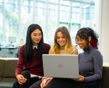 Three students sitting on a couch in the library looking at a laptop screen together.