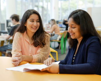 Two students are seated at a yellow table in the library, reviewing printed notes and studying together in a bright, open space.