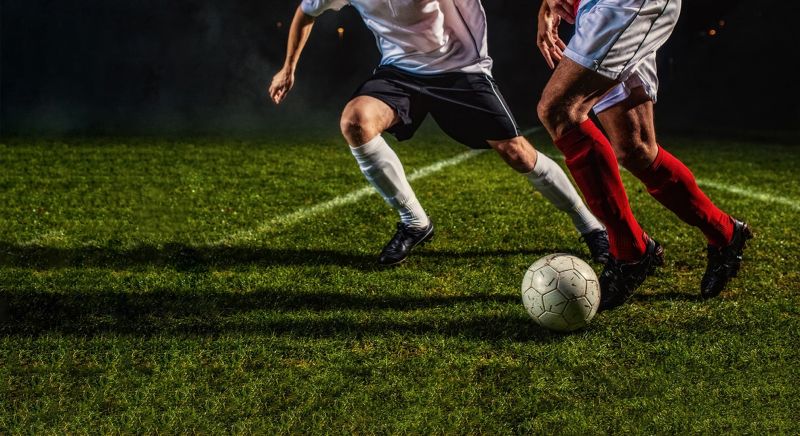 Two soccer players from opposing teams playing on grass field, at night, lit by floodlights