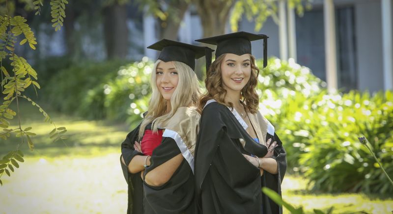 Left to right: sisters Hannah Norris and Lucy Norris standing outside, back-to-back, arms folded and smiling wearing their graduation regalia.