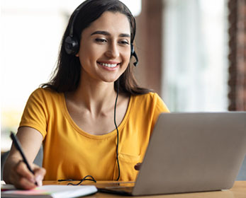 A student wears headphones and takes note attending an online conference on her laptop.