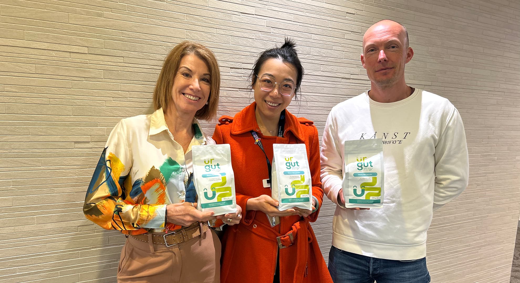 Three people standing against a wall, holding pouches.