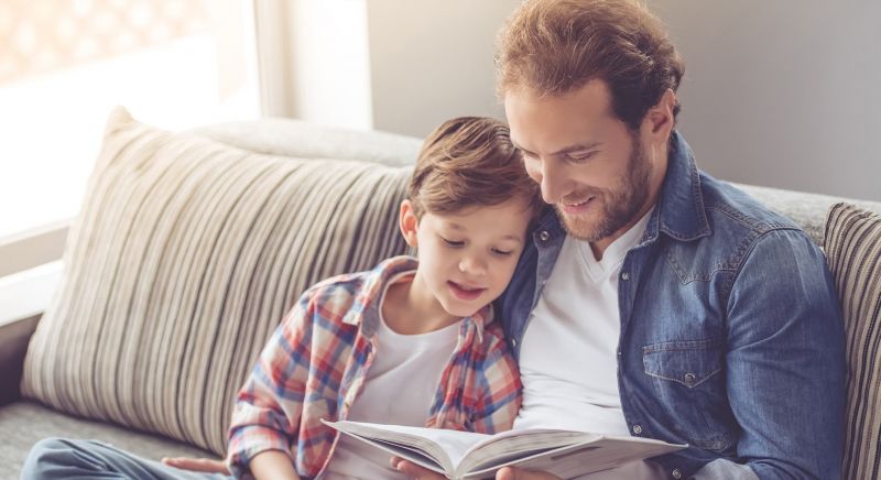 Father and son are reading a book and smiling while spending time together at home