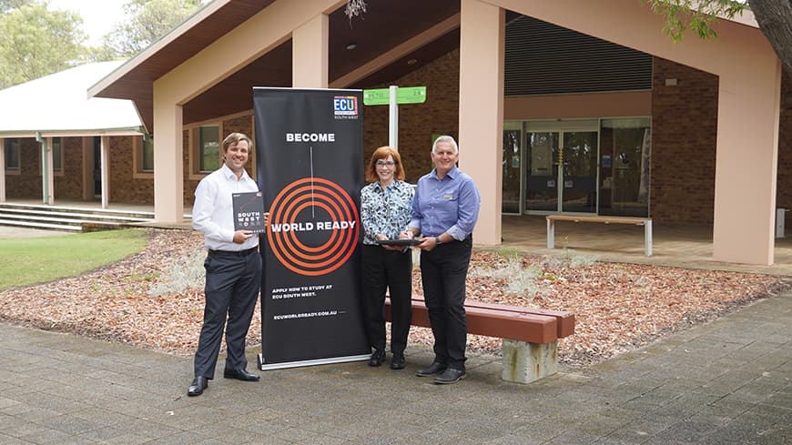 Two men and a woman stand in a bush setting in front of a university branded banner.