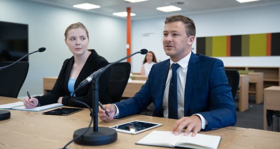 A young woman sits next to a young man behind a desk. They are wearing suits.