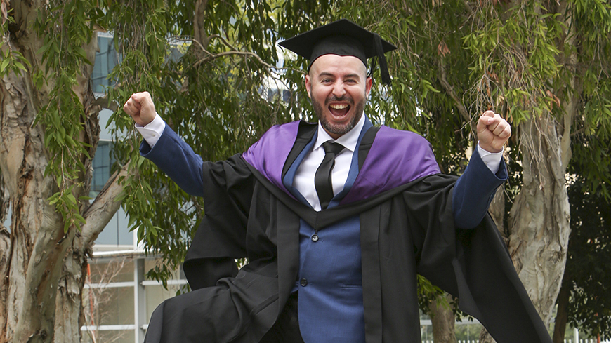 Alvaro Valdes Salazar is dressed in full regalia and cap at his graduation ceremony. He is outdoors and is grinning from ear to ear, he has one knee up in the air and his hands clenched together in triumph.