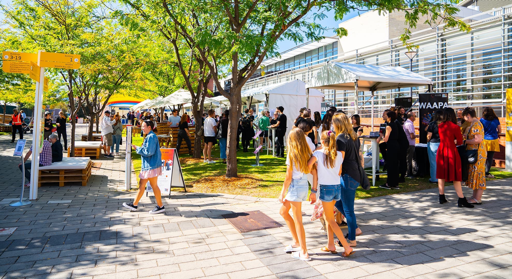 Concrete pathway in foreground, trees in centre, building on right crowds mingling at stalls on grass