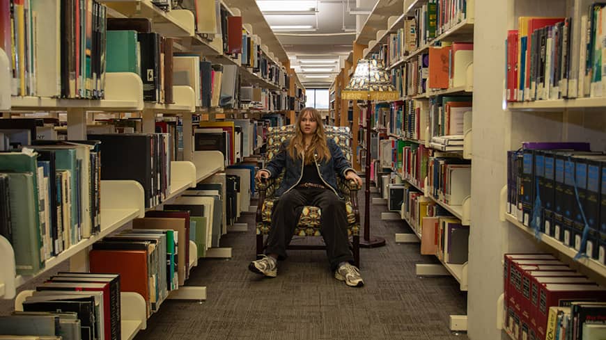 A young adult sitting in an armchair under a lamp in the middle of an aisle in a library, surrounded by books