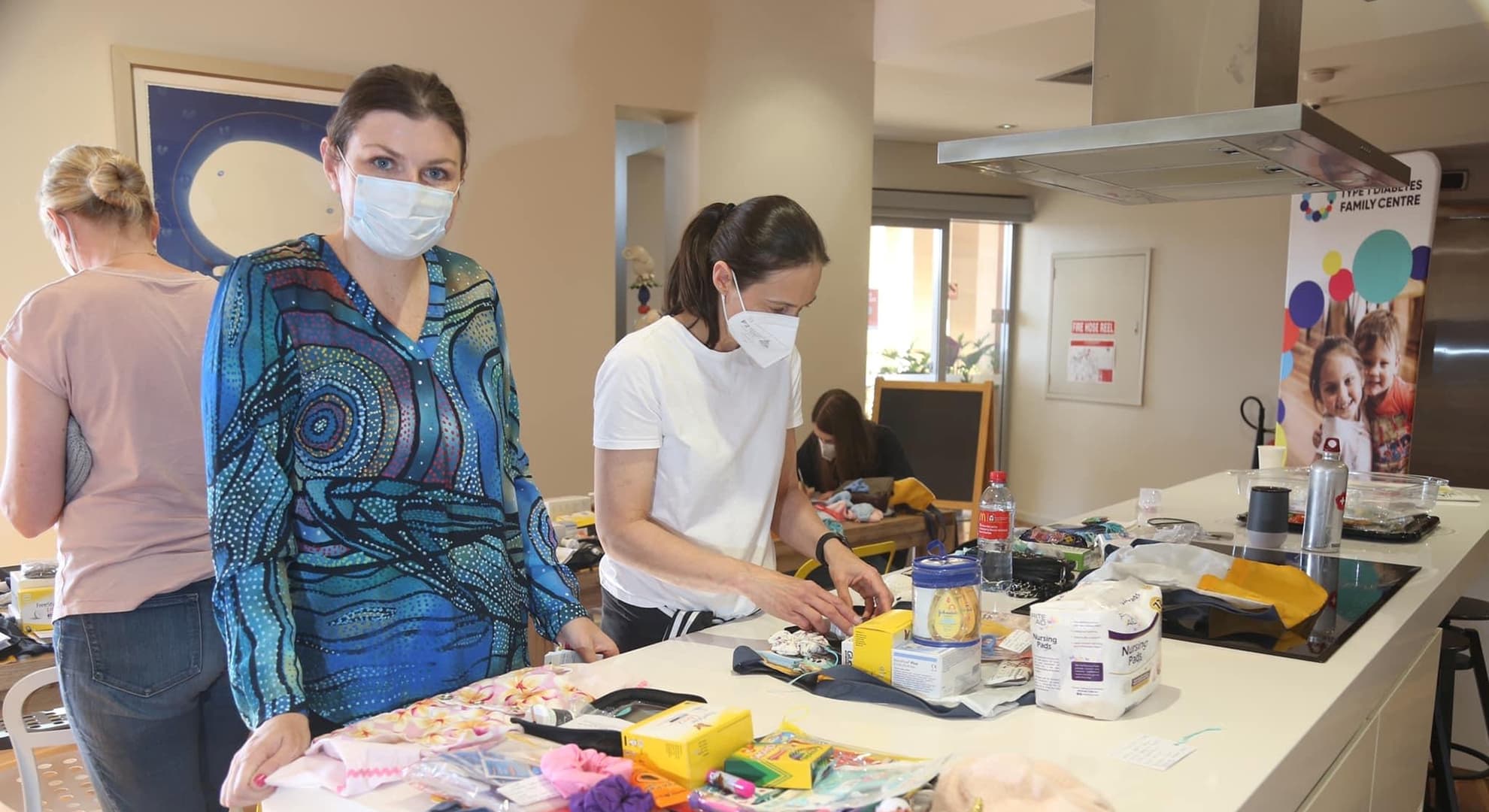 Lady standing at a table looking at camera in COVID face mask, another lady sorting out items on a table