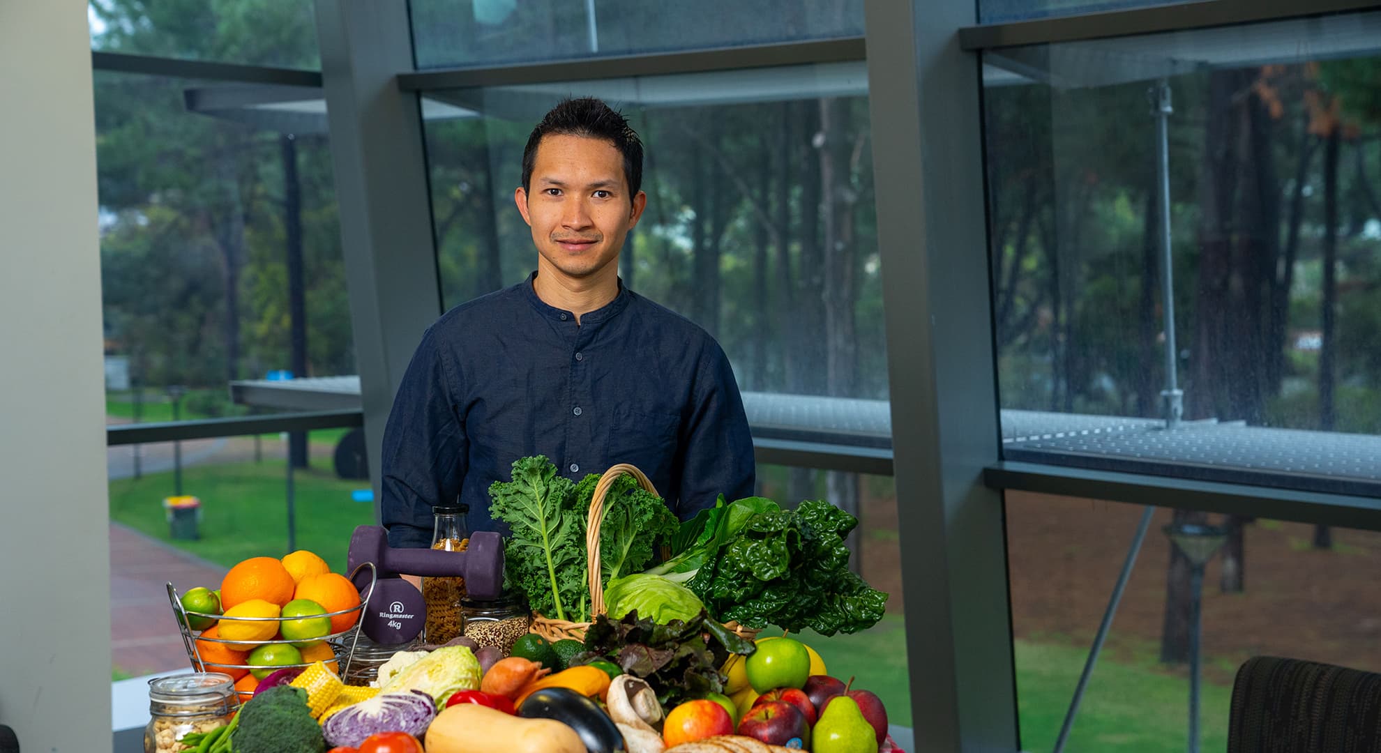 Black haired man standing behind a round table full of fruits and vegetables with red and white check tablecloth