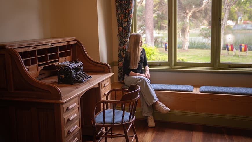 Ayla Lewkowski sits near the window in Edith Cowan House and looks out. There is a wooden desk to the side with a typewriter.