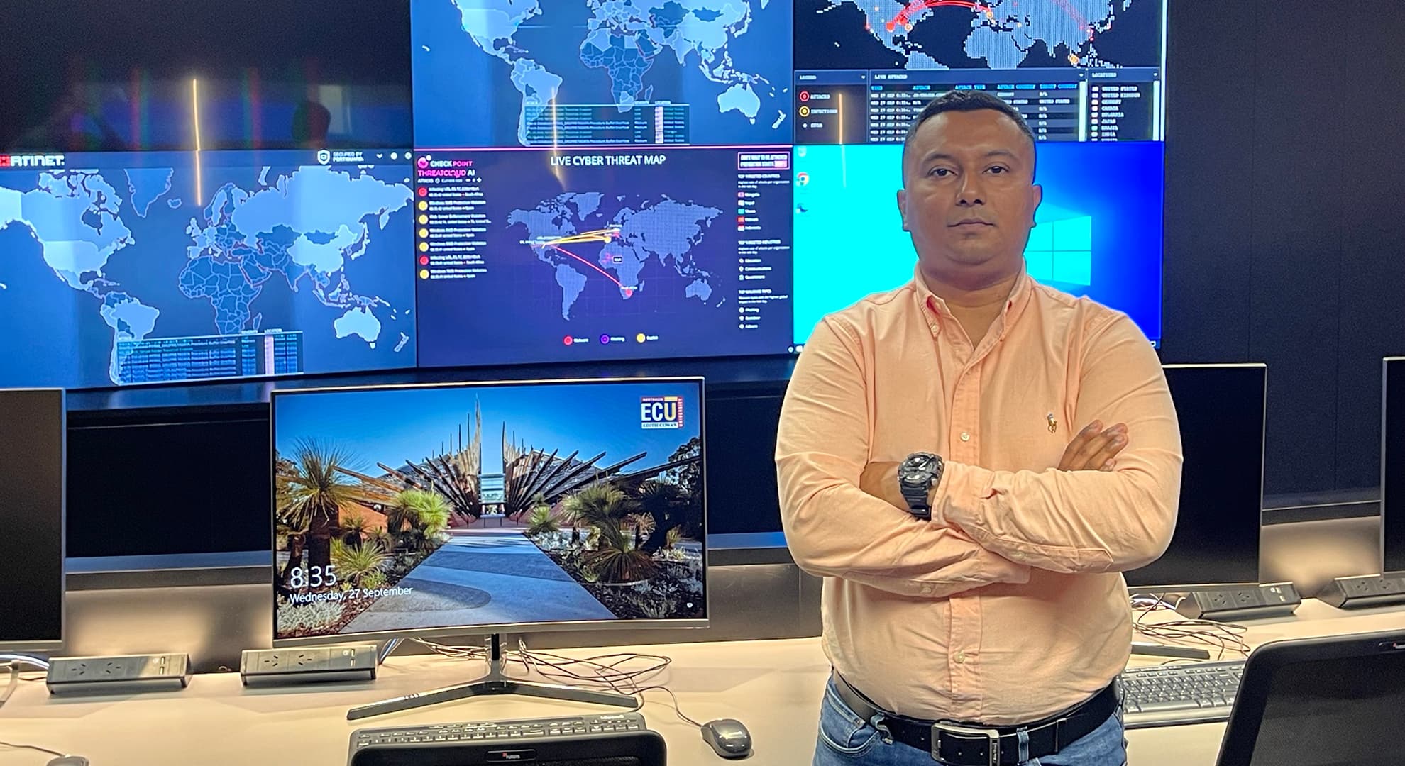 Senior Lecturer of Computing and Security Dr Mohi Ahmed stands in front of computers and monitors lit up with live cyber threat maps.