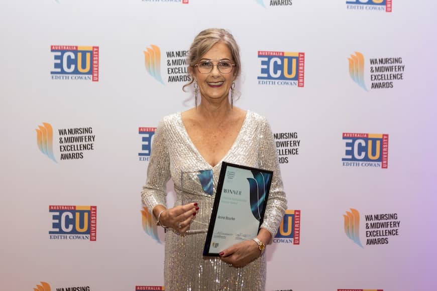 Anne Bourke wears a sequined silver dress, dangly earrings and silver framed glasses. She smiles and holds out her framed award and glass trophy.