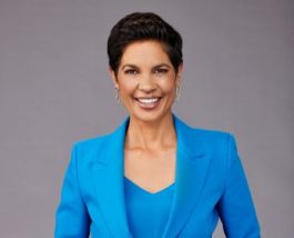Narelda Jacobs, wearing a blue suit in front of a grey background, smiling to camera