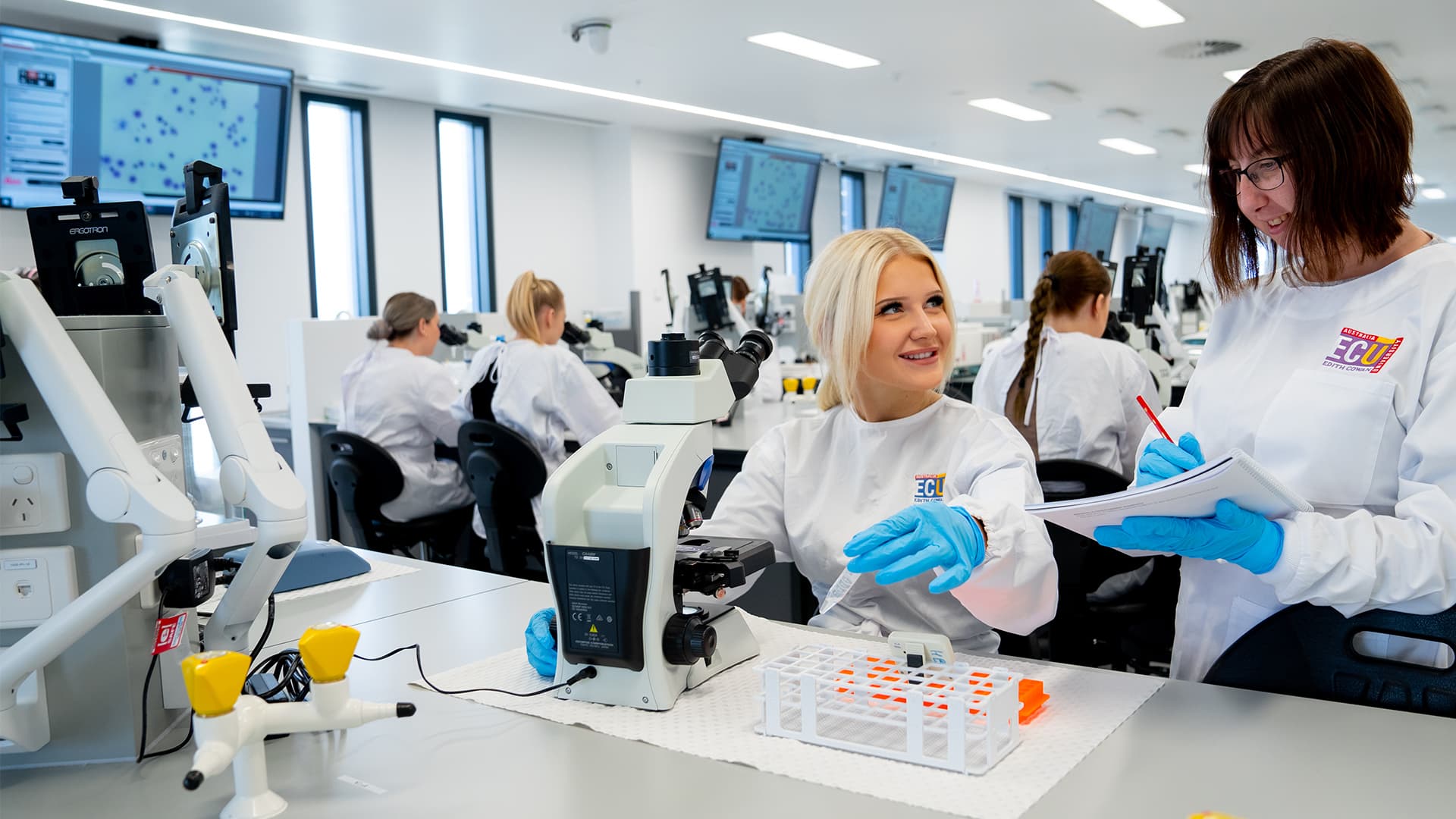 Two students in a laboratory at a desk with test tubes and a microscope.