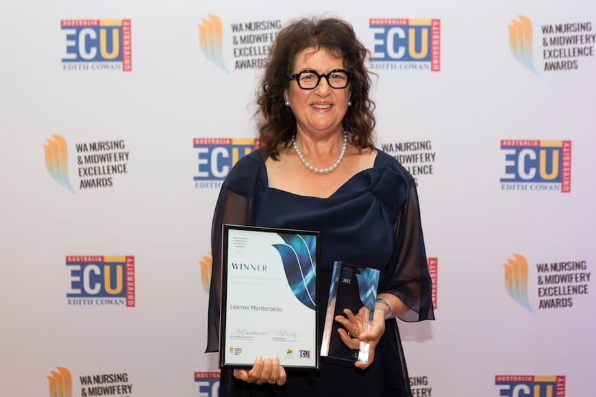 Leanne Monterosso wears a navy gown with a pearl necklace and black round glasses. She smiles and holds out her framed award and glass trophy.