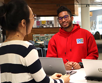 A student who is employed as a Library Peer Assistant is smiling at the camera, stationed at Joondalup Campus Library. She wears the red jumper that is the peers’ uniform.