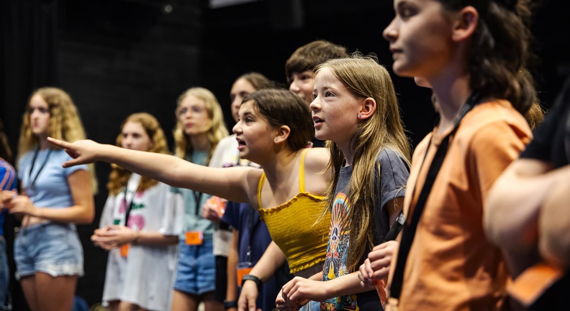 children in standing a semi-circle facing at right angles to the camera, two girls in the centre in focus one pointing with yellow elastic top with shoe string straps