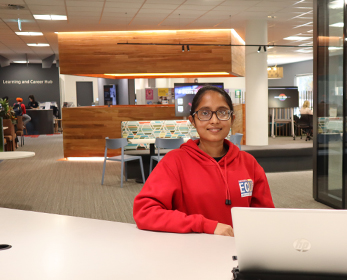 A Library Peer Assistant wears the red hoodie that is the peer uniform. She is seated at the welcome desk at the Joondalup Library's Learning and Career Hub.