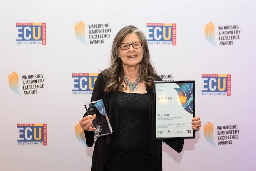 Anne Watts wears a black gown with a intricate blue necklackce and red glasses, she holds out a framed award and a glass trophy and smiles.