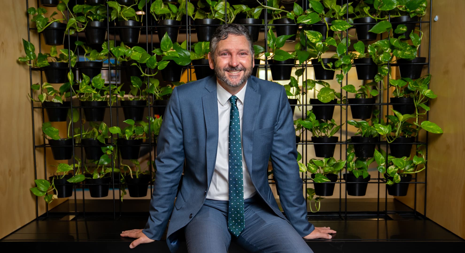 Professor with a beard in suit sitting on black bench in front of wall of small potted greenery