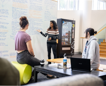 Three students collaborate in a Library study space with a whiteboard, vending machine, and natural light from nearby windows.