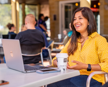 Student sitting at an outdoor campus table, using a laptop with a coffee and phone nearby, smiling while studying in a relaxed, social environment with other people in the background.