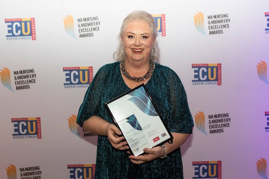 Jodie Atkinson wears a metallic teal-coloured dress with a chunky bead necklace, she holds her framed award and smiles.