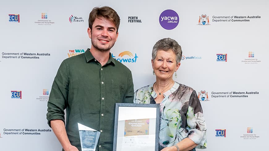 Cameron Carr and Estelle Dragun who are dressed up in smart casual clothes, smile and hold the Charmaine Dragun Memorial Award.