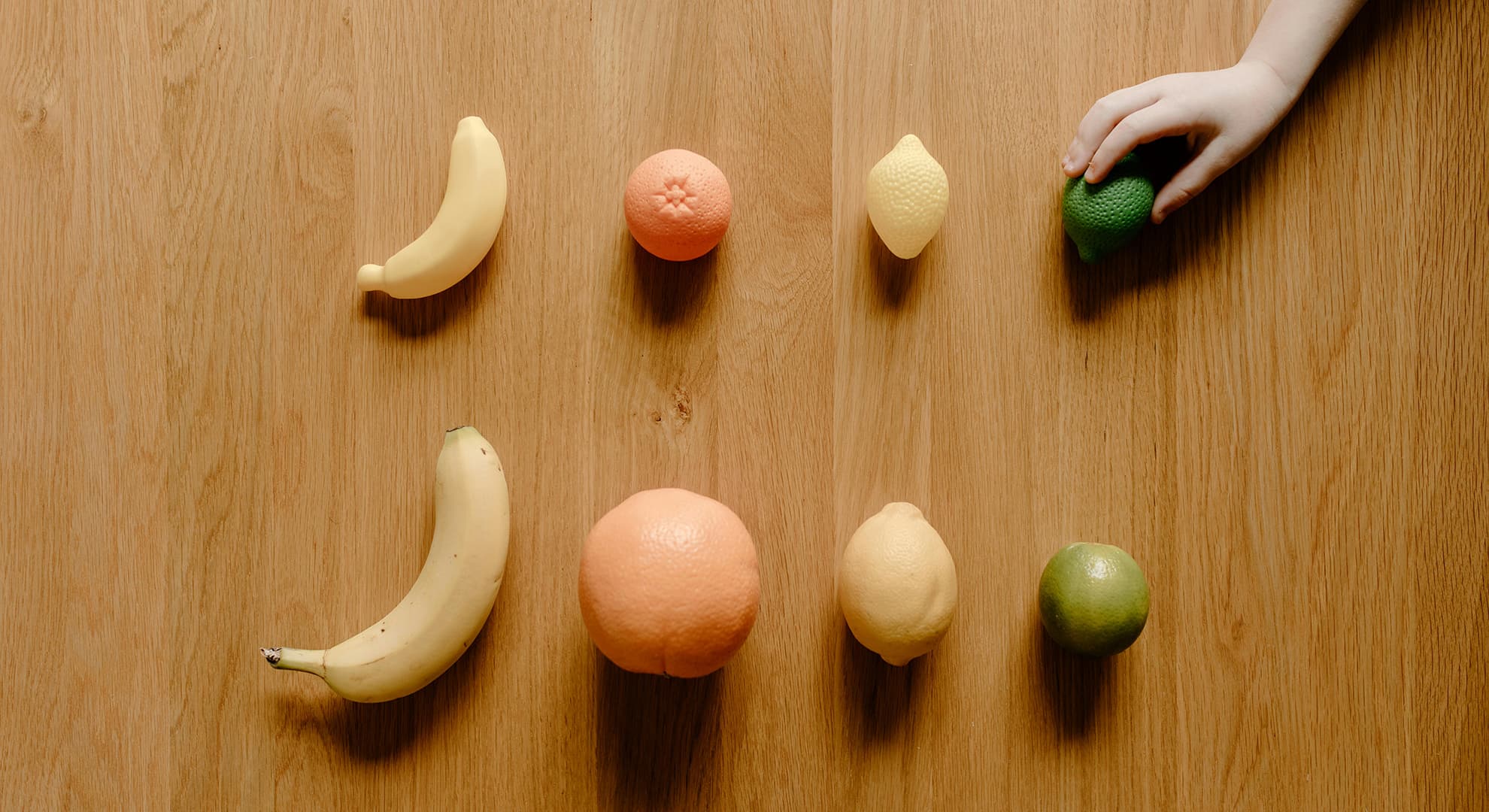 Plastic fruit and real fruit lined up, with child's hand.