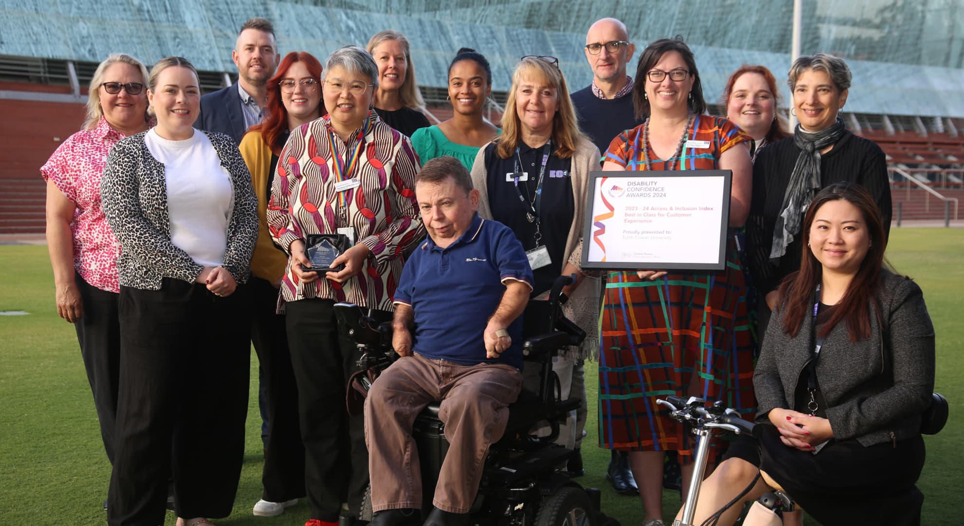 Staff group of 14 people standing outside on lawn, man in wheelchair in centre foreground and lady sitting on scooter with prosthetic leg at far right, framed certificate being held up in the middle of seated staff