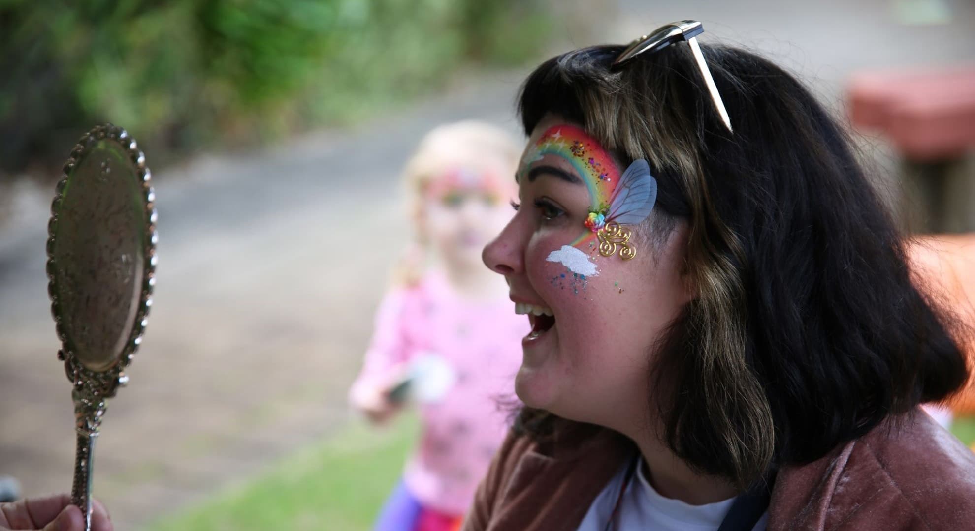 young woman in profile, rainbow with sparkles painted on her face looking into a small gold mirror being held up to show her